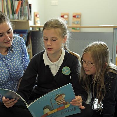 A teacher reading a picture book with two pupils on the floor of the classroom library