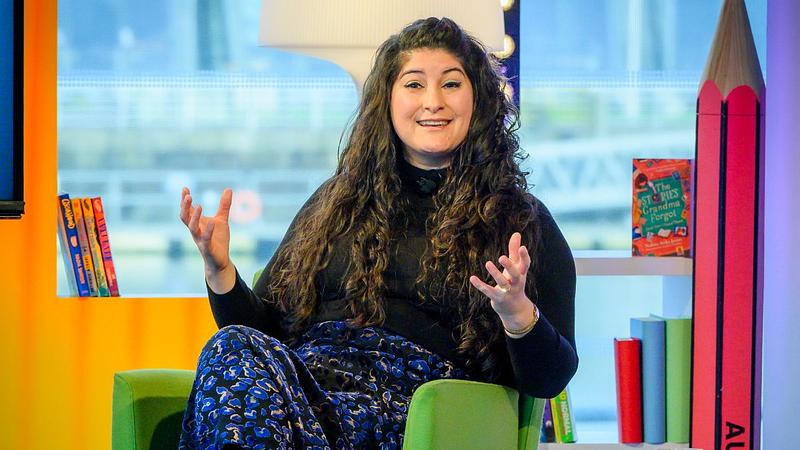 A smiling woman sits in a colourful TV studio with an oversized pencil and books behind her